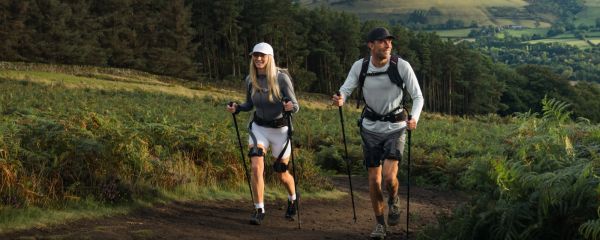 A young man and woman are hiking together, walking up a steady hill on a sunny afternoon. They are smiling as the sun shines towards them. Behind them is a view of lush green tumbling hills and rolling countryside. Both are wearing Ecoskeletons